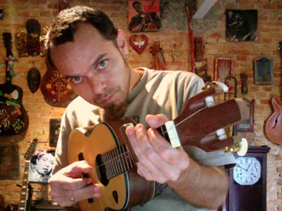 Eduardo Sardinha tocando violão em ambiente interno com parede de tijolos e vários instrumentos de corda ao fundo