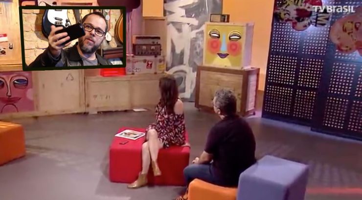 Estúdio colorido da TV Brasil com duas pessoas sentadas e Eduardo Sardinha em uma tela segurando um smartphone