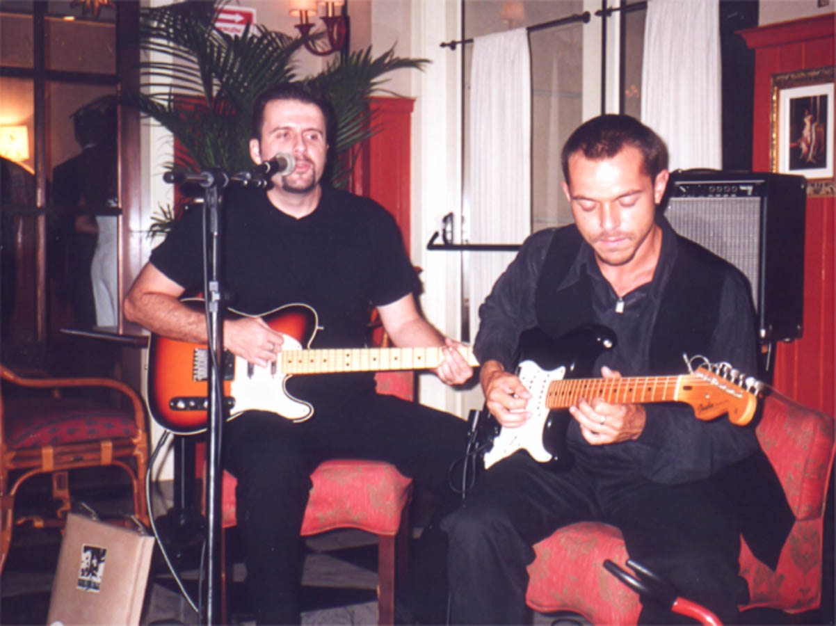 Eduardo Sardinha e Luiz Carlos Big, integrantes do Bluescake, tocam guitarras elétricas em apresentação musical. Elementos visíveis: duo, guitarristas, microfone, amplificador, cabos, poltronas vermelhas, instrumentos de cordas e ambiente interno. Arquivo: Eduardo Sardinha / Sardinha17.