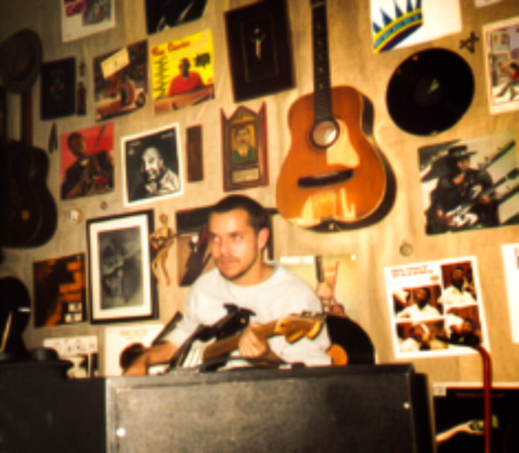 Integrante do Bluescake sentado com guitarra em ambiente dedicado à música. Elementos visíveis: músico, guitarra, amplificador, violões na parede, disco de vinil, cartazes, fotografias, molduras, luminária e coleção musical. Bluescake: Eduardo Sardinha e Luiz Carlos Big. Arquivo: Eduardo Sardinha / Sardinha17.
