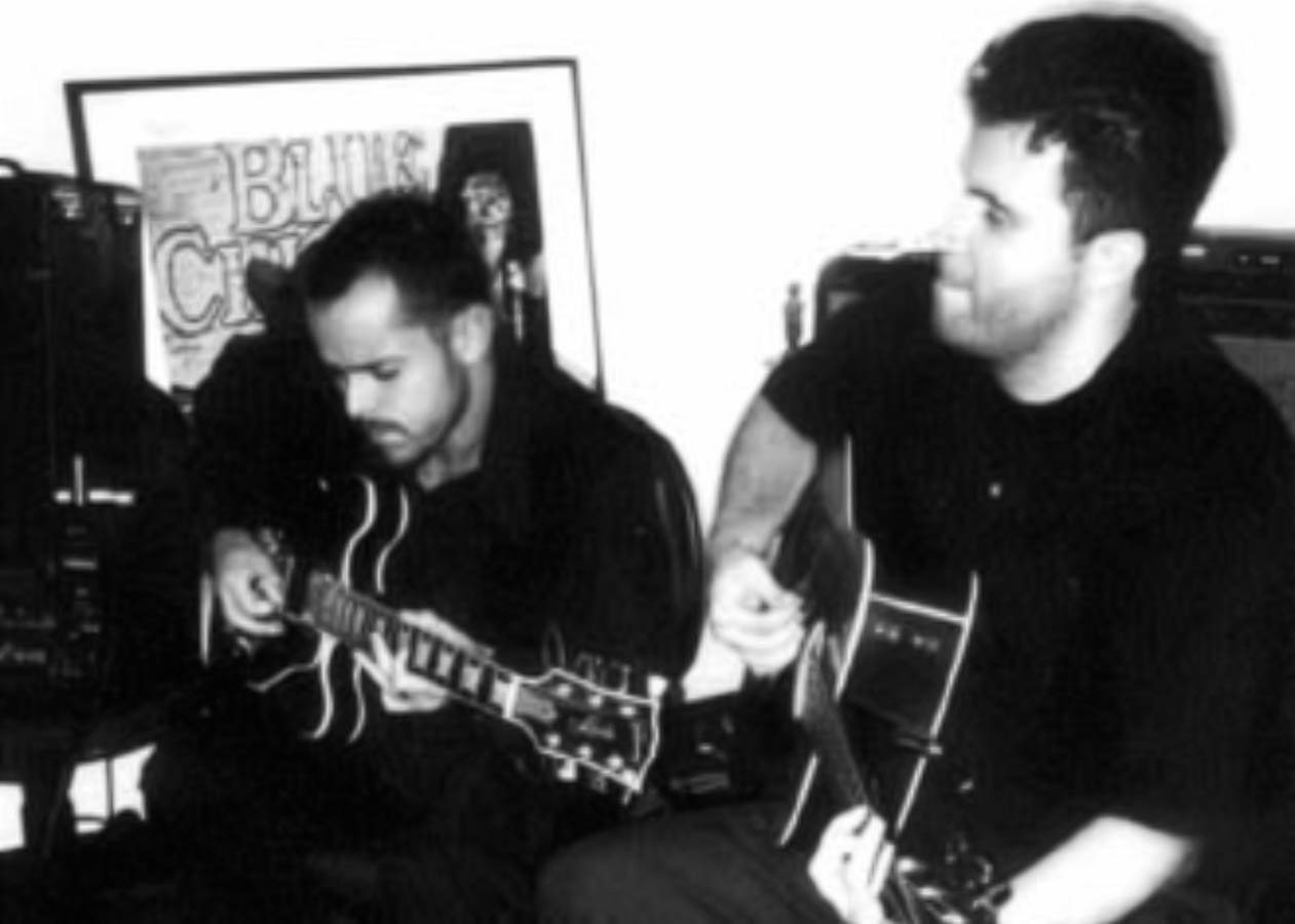 Retrato preto e branco do Bluescake com Eduardo Sardinha e Luiz Carlos Big tocando guitarra semiacústica e violão. Elementos visíveis: duo, músicos, instrumentos de cordas, amplificador, cartaz, mãos, alto contraste e memória musical. Arquivo: Eduardo Sardinha / Sardinha17.