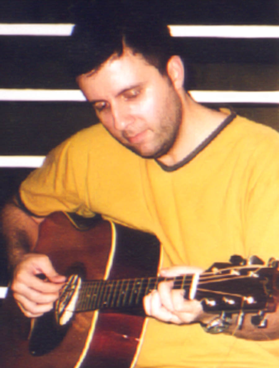Retrato vertical de integrante do Bluescake tocando violão acústico. Elementos visíveis: músico, violonista, violão, cordas, braço do instrumento, mãos, madeira, camiseta amarela e ambiente interno. Bluescake: Eduardo Sardinha e Luiz Carlos Big. Arquivo: Eduardo Sardinha / Sardinha17.