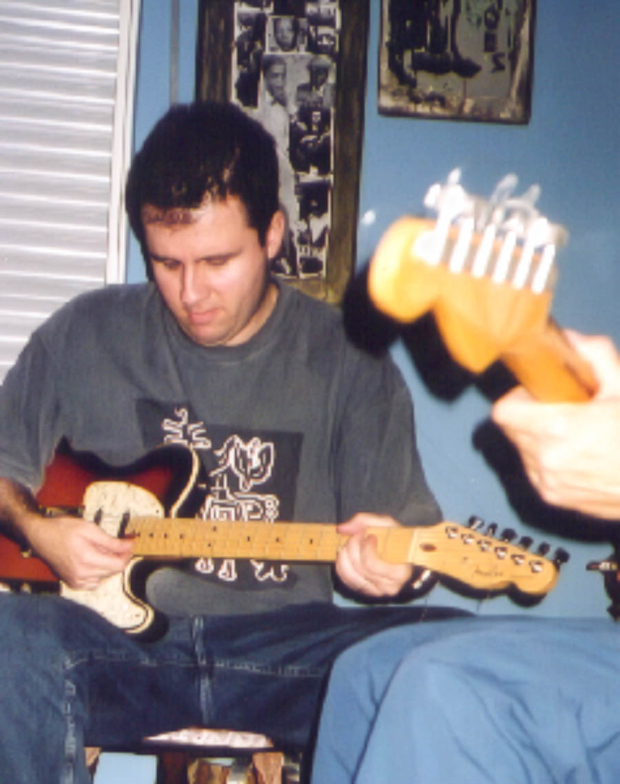 Integrante do Bluescake toca guitarra elétrica em ambiente decorado com fotografias e objetos musicais; outro braço de guitarra aparece em primeiro plano. Elementos visíveis: guitarra Telecaster, captadores, mãos, banco, parede azul, molduras e perspectiva. Bluescake: Eduardo Sardinha e Luiz Carlos Big. Arquivo: Eduardo Sardinha / Sardinha17.