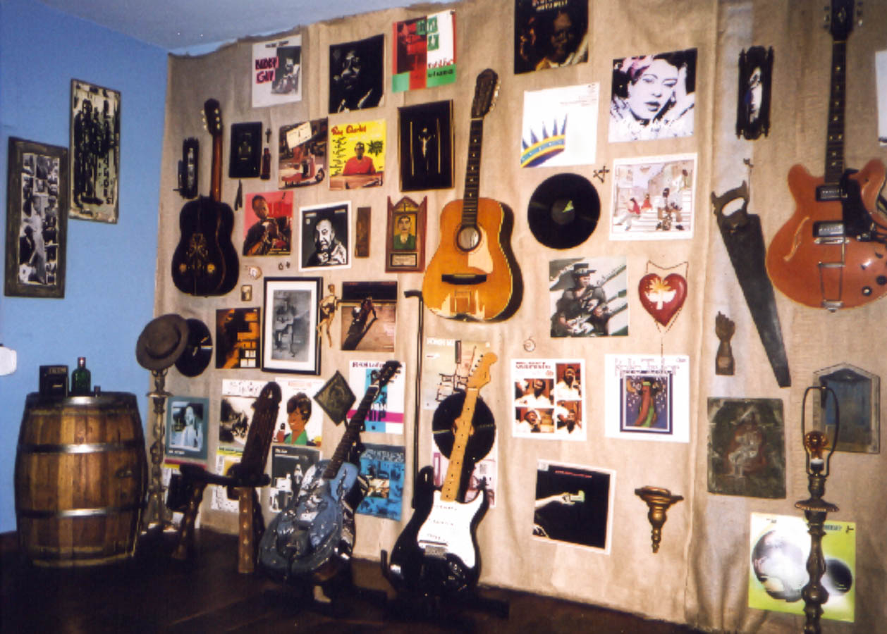 Interior ligado ao universo do Bluescake, com coleção de instrumentos e memória musical. Elementos visíveis: guitarras elétricas, violões, discos de vinil, capas, fotografias, cartazes, chapéus, molduras, barril de madeira, luminária e parede azul. Bluescake: Eduardo Sardinha e Luiz Carlos Big. Arquivo: Eduardo Sardinha / Sardinha17.