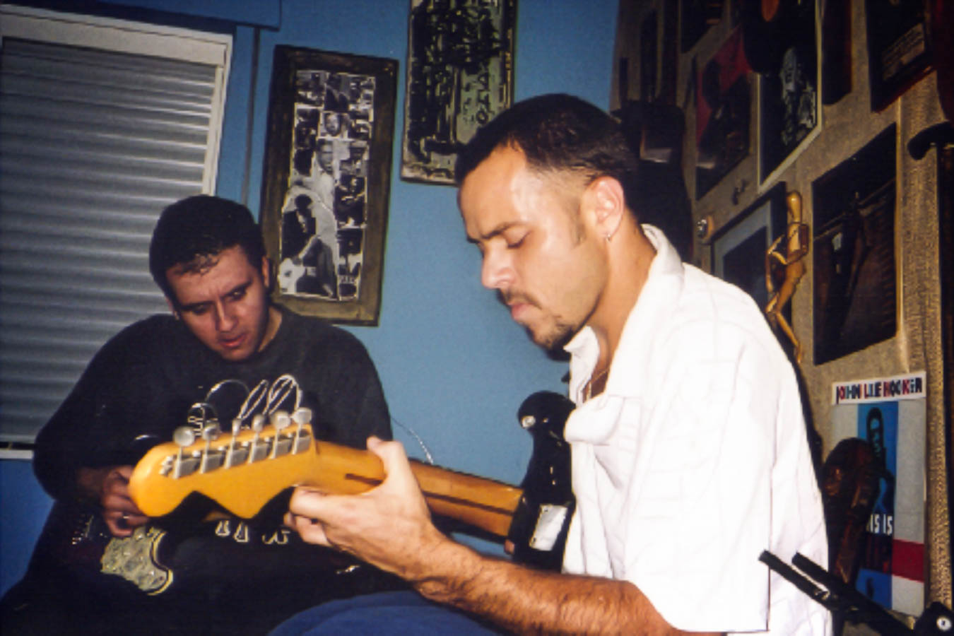 Eduardo Sardinha e Luiz Carlos Big tocam guitarras elétricas em um ensaio do Bluescake. Elementos visíveis: duo, músicos, guitarras, mãos, banco, parede azul, fotografias, cartazes, instrumentos, interior e luz direta. Arquivo: Eduardo Sardinha / Sardinha17.