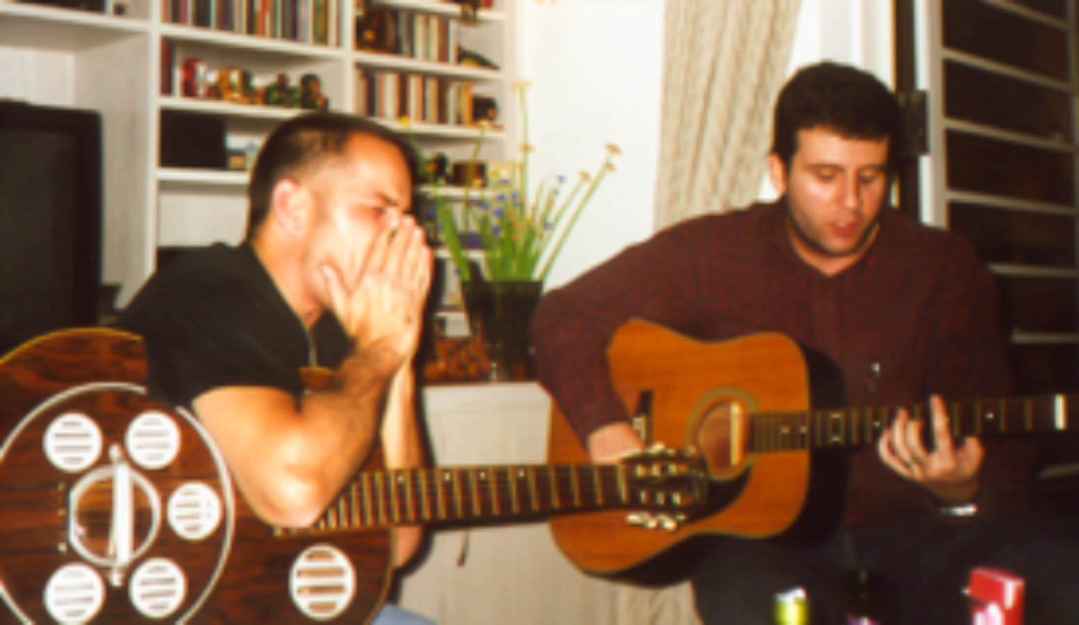 Eduardo Sardinha e Luiz Carlos Big em encontro do Bluescake, com um integrante tocando gaita e o outro violão acústico. Elementos visíveis: duo, gaita, violões, mãos, instrumentos de cordas, estante com livros, janela, planta e ambiente doméstico. Arquivo: Eduardo Sardinha / Sardinha17.