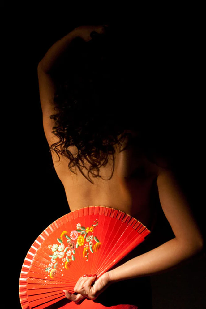 Retrato de costas de Karla Jacobina, com braço elevado, gesto coreográfico e leque vermelho floral contra fundo preto. Fotografia autoral de Eduardo Sardinha — Sardinha17. Palavras-chave: dança, leque, vermelho, figurino, expressão corporal, sombras, retrato.