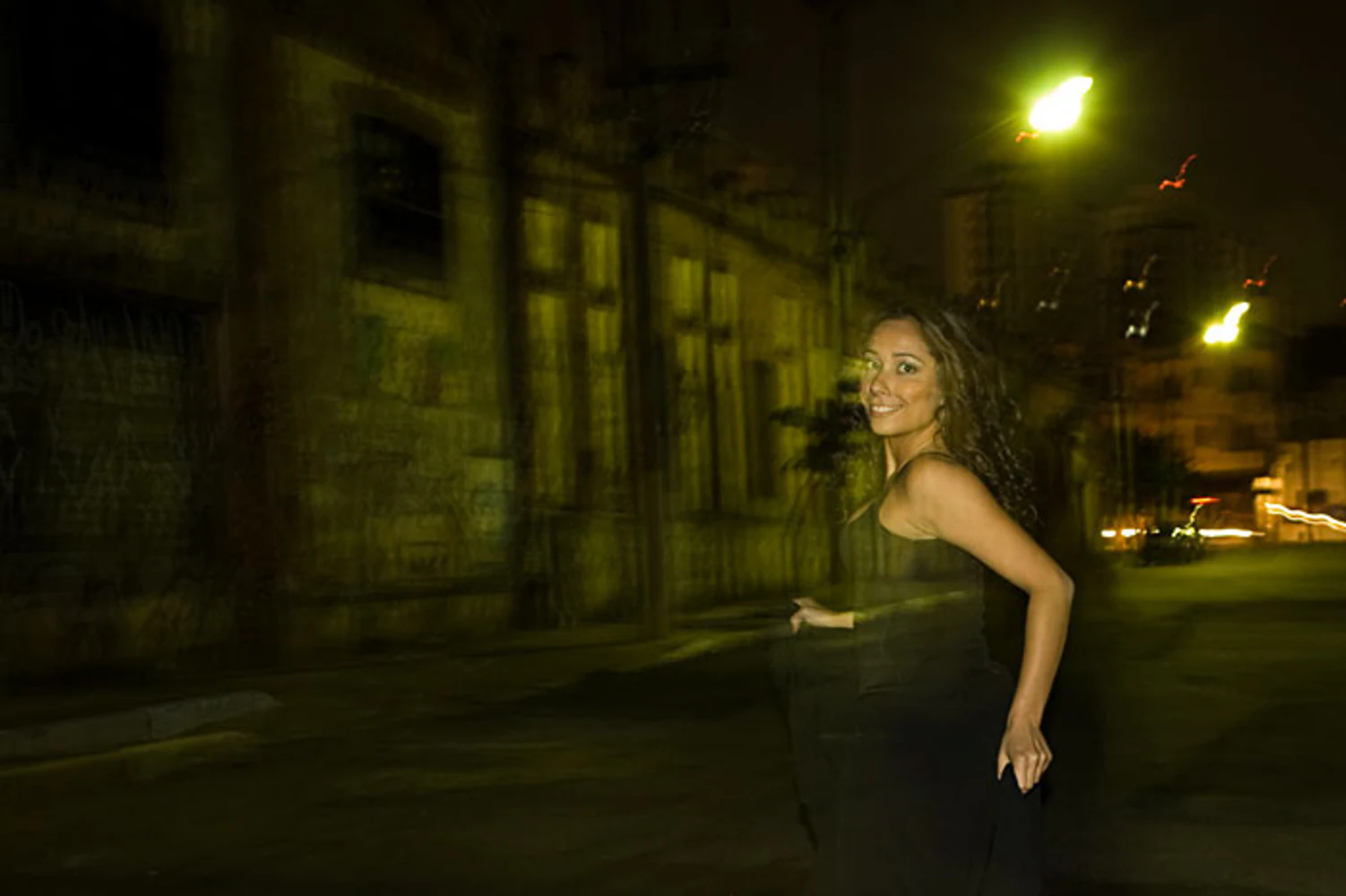 Retrato urbano noturno de Karla Jacobina em movimento, com fachada arquitetônica, rua vazia, iluminação amarela e efeito de longa exposição. Fotografia autoral de Eduardo Sardinha — Sardinha17. Palavras-chave: cidade à noite, arquitetura, rua, movimento, luz amarela, longa exposição.