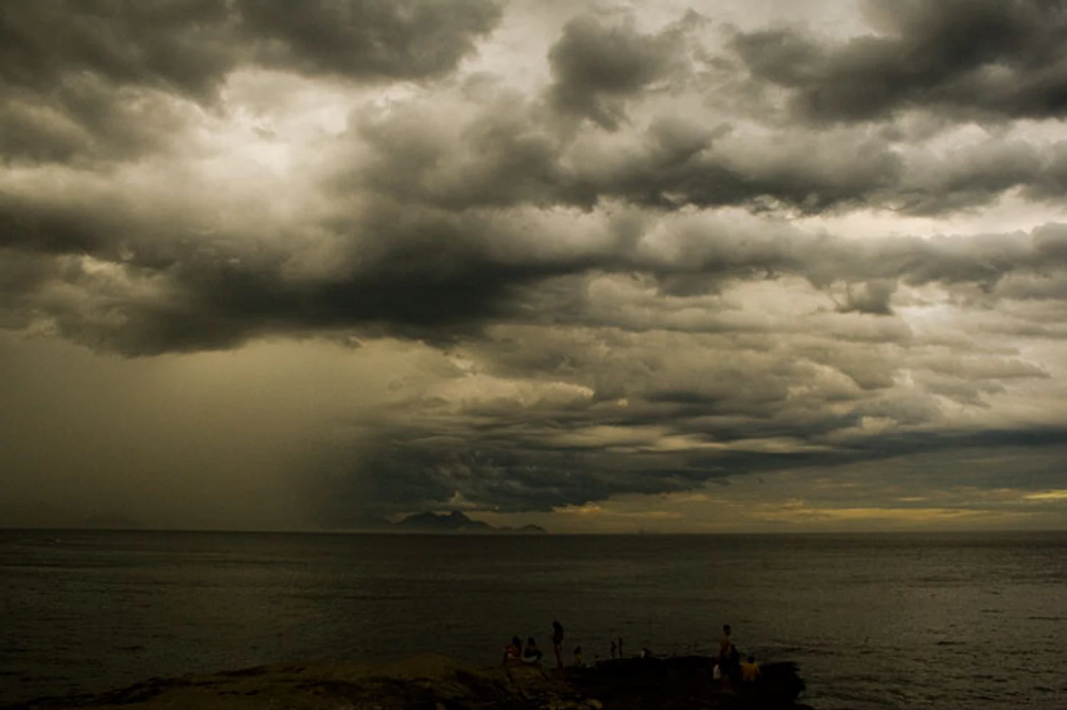 Nuvens densas de tempestade sobre o mar, com chuva no horizonte e pessoas sobre as rochas
