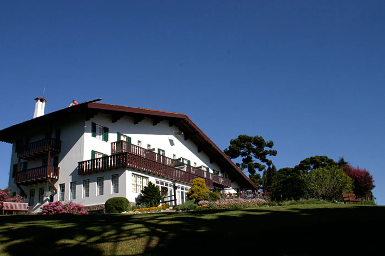 Vista externa do Hotel Toriba, em Campos do Jordão, com arquitetura de inspiração alpina, fachada branca, telhado inclinado, varandas de madeira, jardim e araucárias. Fotografia autoral de Eduardo Sardinha — Sardinha17. Palavras-chave: arquitetura, hotel, fachada, Serra da Mantiqueira, turismo.