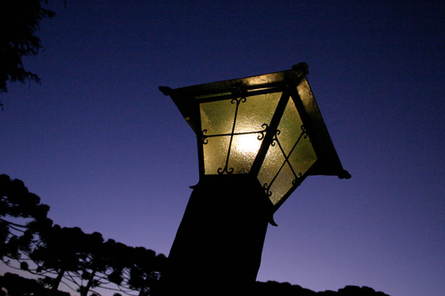 Luminária de jardim em ferro e vidro acesa ao anoitecer, fotografada em contraplongée contra céu azul profundo e araucárias em silhueta, no Hotel Toriba. Fotografia autoral de Eduardo Sardinha — Sardinha17. Palavras-chave: luminária, luz, noite, arquitetura, detalhe, perspectiva.