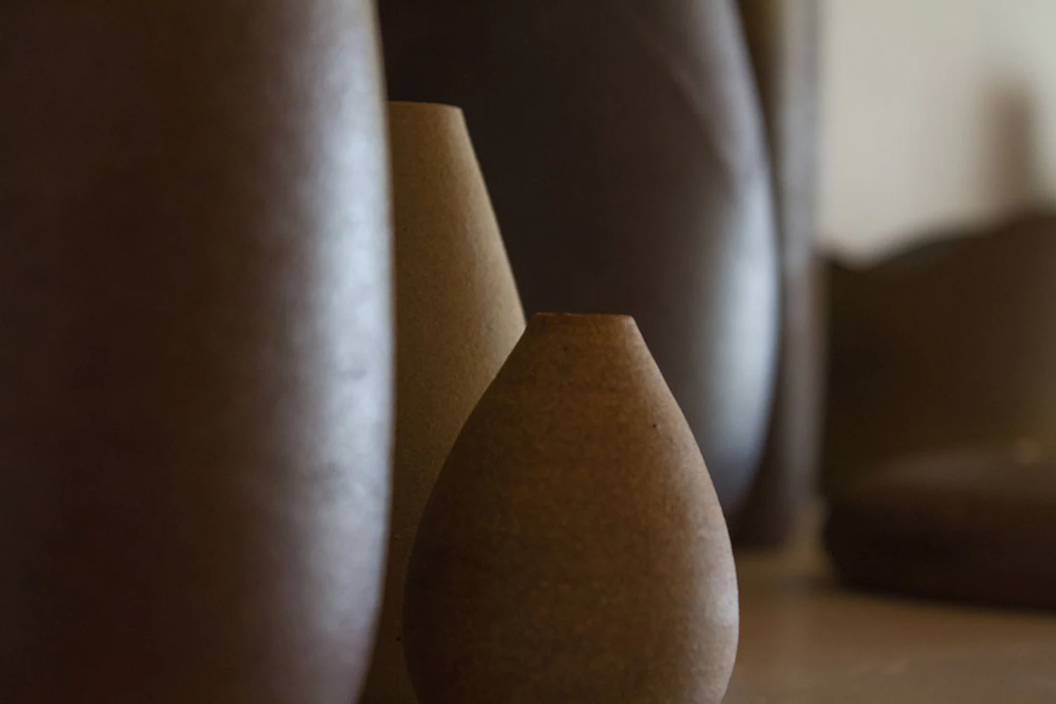 Composição editorial com vasos e recipientes cerâmicos de Kimi Nii em tons terrosos. Elementos visíveis: cerâmica de alta temperatura, vasos, formas orgânicas, superfície fosca, marrom, bege, luz lateral, sombra e profundidade de campo. Fotografia autoral de Eduardo Sardinha / Sardinha17 para projeto Eurobike. Contexto: ateliê Kimi Nii, São Paulo, SP, Brasil.