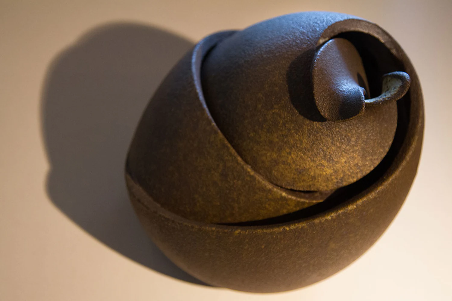 Escultura cerâmica de Kimi Nii formada por volumes curvos encaixados e concêntricos. Elementos visíveis: arte contemporânea, cerâmica de alta temperatura, escultura, geometria orgânica, curva, espiral, marrom, textura, luz dirigida e sombra projetada. Fotografia autoral de Eduardo Sardinha / Sardinha17 para a Eurobike. Contexto: ateliê Kimi Nii, São Paulo, SP, Brasil.
