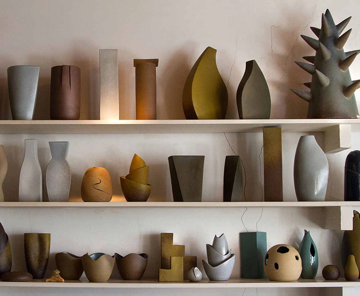 Vista ampla de coleção de Kimi Nii com vasos, esculturas e objetos utilitários organizados em prateleiras. Elementos visíveis: cerâmica artesanal, formas geométricas e orgânicas, vasos, recipientes, esculturas, repetição, branco, cinza, verde, ocre, marrom e ambiente de ateliê. Fotografia autoral de Eduardo Sardinha / Sardinha17 para a Eurobike. Local: São Paulo, SP, Brasil.