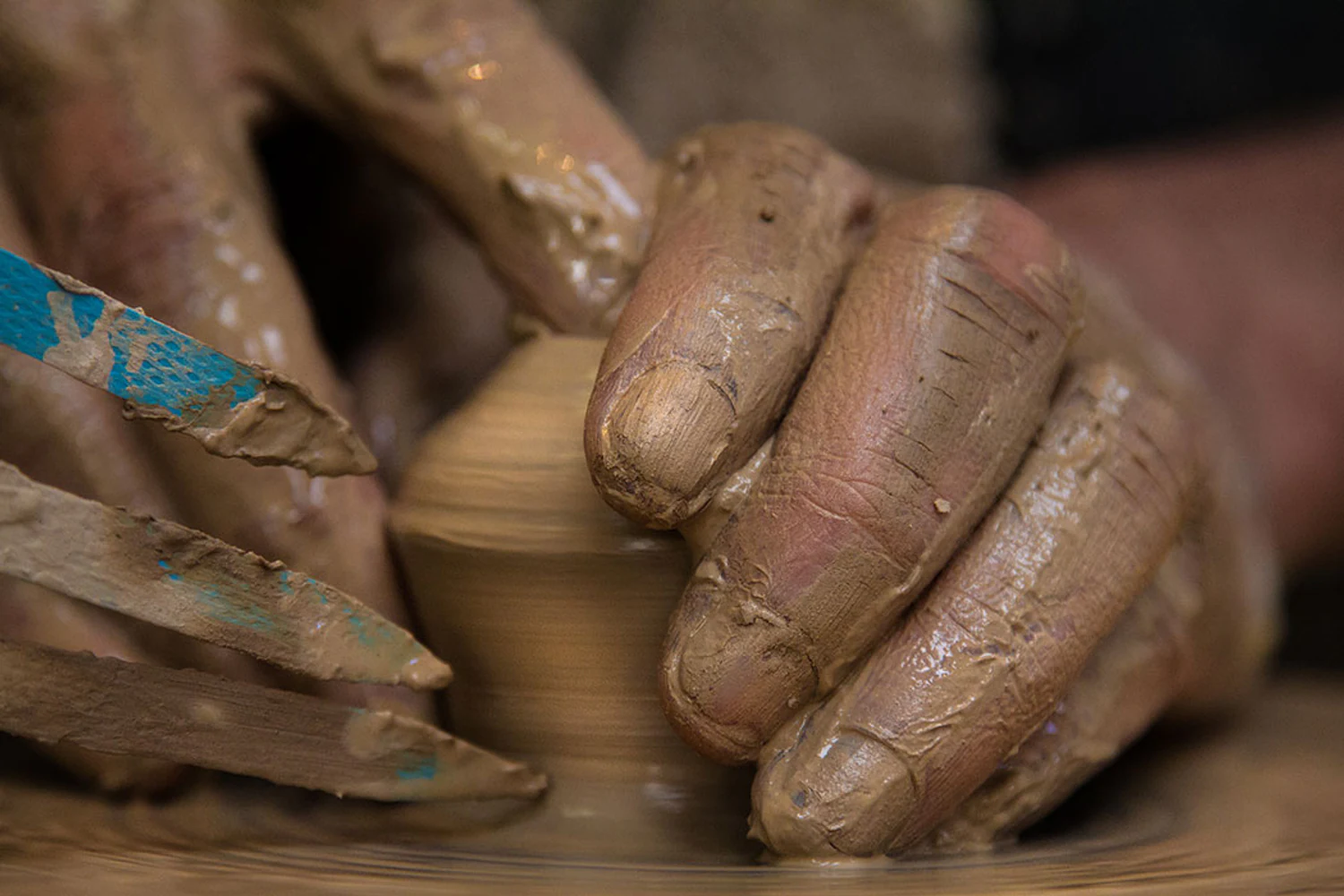 Close do processo artesanal com mãos cobertas de argila modelando uma peça no torno em movimento. Elementos visíveis: mãos, dedos, barro, argila, torno de cerâmica, ferramenta, modelagem, textura, gesto, artesanato e trabalho manual. Fotografia autoral de Eduardo Sardinha / Sardinha17 para a Eurobike. Contexto: ateliê Kimi Nii, São Paulo, SP, Brasil.