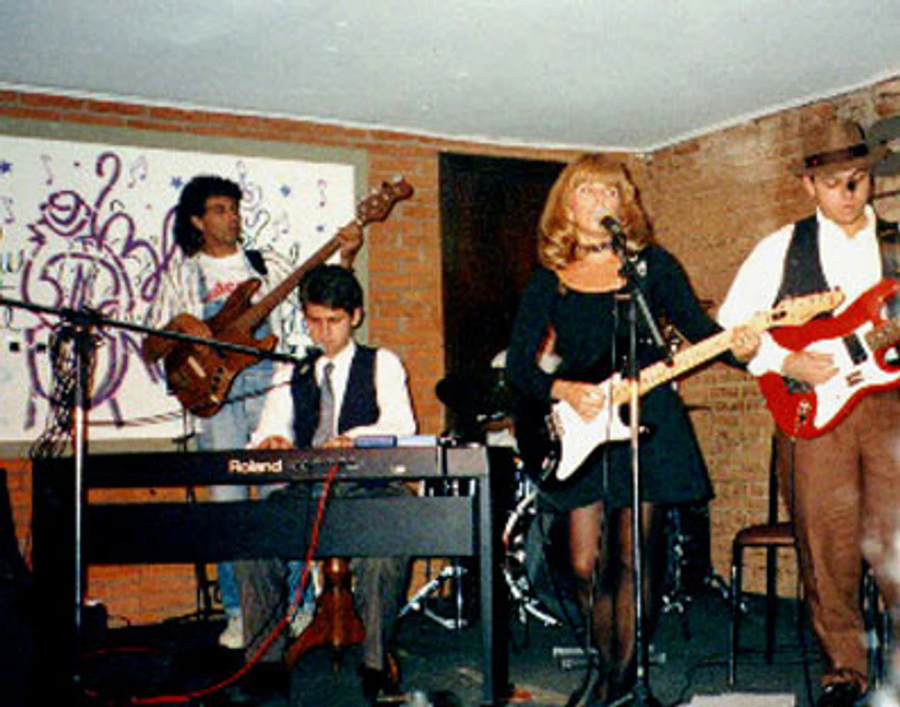 Apresentação ao vivo da Mississampa Blues Band com Marina Teixeira de Mello, Eduardo Sardinha e Rogério Mello, acompanhados por músicos não identificados. Elementos visíveis: cantora, guitarra elétrica, teclado Roland, contrabaixo, bateria, microfones, palco, tijolos aparentes, painel gráfico, performance e blues. Fotografia e arquivo autoral de Eduardo Sardinha / Sardinha17.