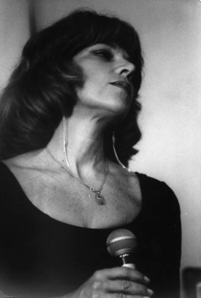 Retrato em preto e branco de Marina Teixeira de Mello, mãe de Eduardo Sardinha e Rogério Mello e integrante da Mississampa Blues Band, segurando um microfone. Elementos visíveis: cantora, vocalista, microfone, colar, vestido escuro, close, música e retrato feminino. Fotografia e arquivo autoral de Eduardo Sardinha / Sardinha17.