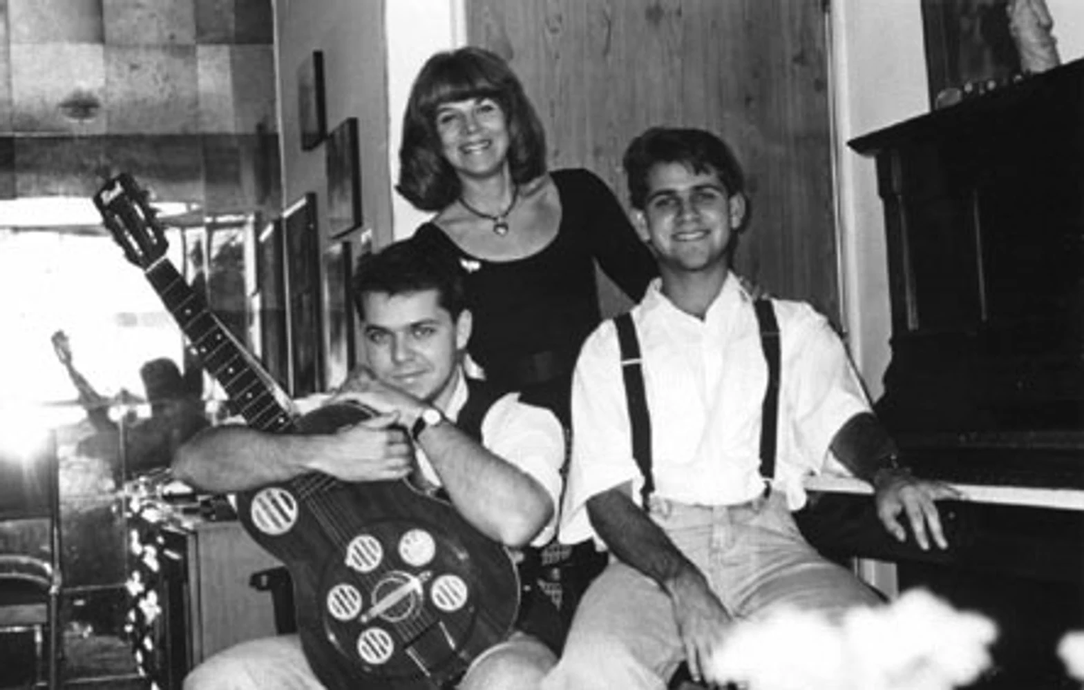Retrato familiar e musical da Mississampa Blues Band com Marina Teixeira de Mello em pé ao centro, Eduardo Sardinha sentado à esquerda com violão e Rogério Mello à direita junto ao piano. Elementos visíveis: mãe e filhos, músicos, violão, piano, suspensórios, madeira, espelho, mobiliário e ambiente doméstico. Fotografia e arquivo autoral de Eduardo Sardinha / Sardinha17.